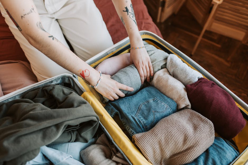 A person is packing a large, open suitcase filled with neatly folded clothing items, including jeans, sweaters, and socks, inside a residential room with wooden flooring. The individual, wearing a whi