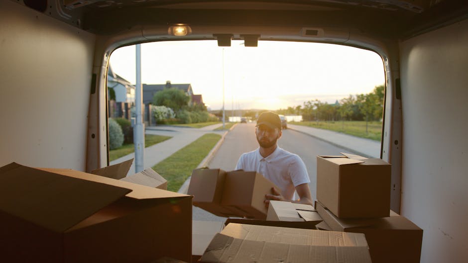 A person loading cardboard boxes into the back of a moving van during a home relocation. The photograph is taken from inside the van, showing the interior with some boxes arranged on the floor and par