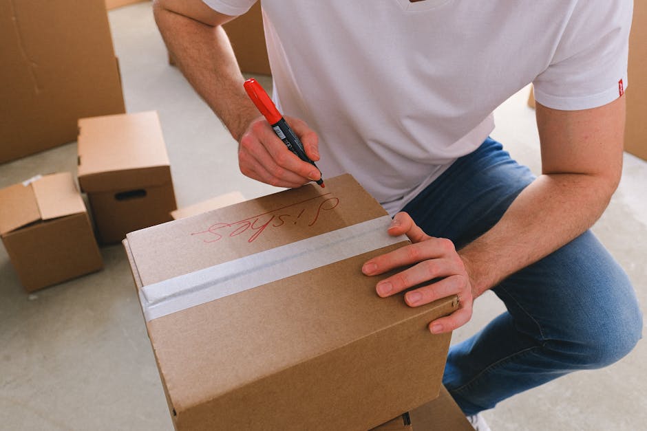 A person loading cardboard boxes into the back of a moving van during a home relocation. The photograph is taken from inside the van, showing the interior with some boxes arranged on the floor and par